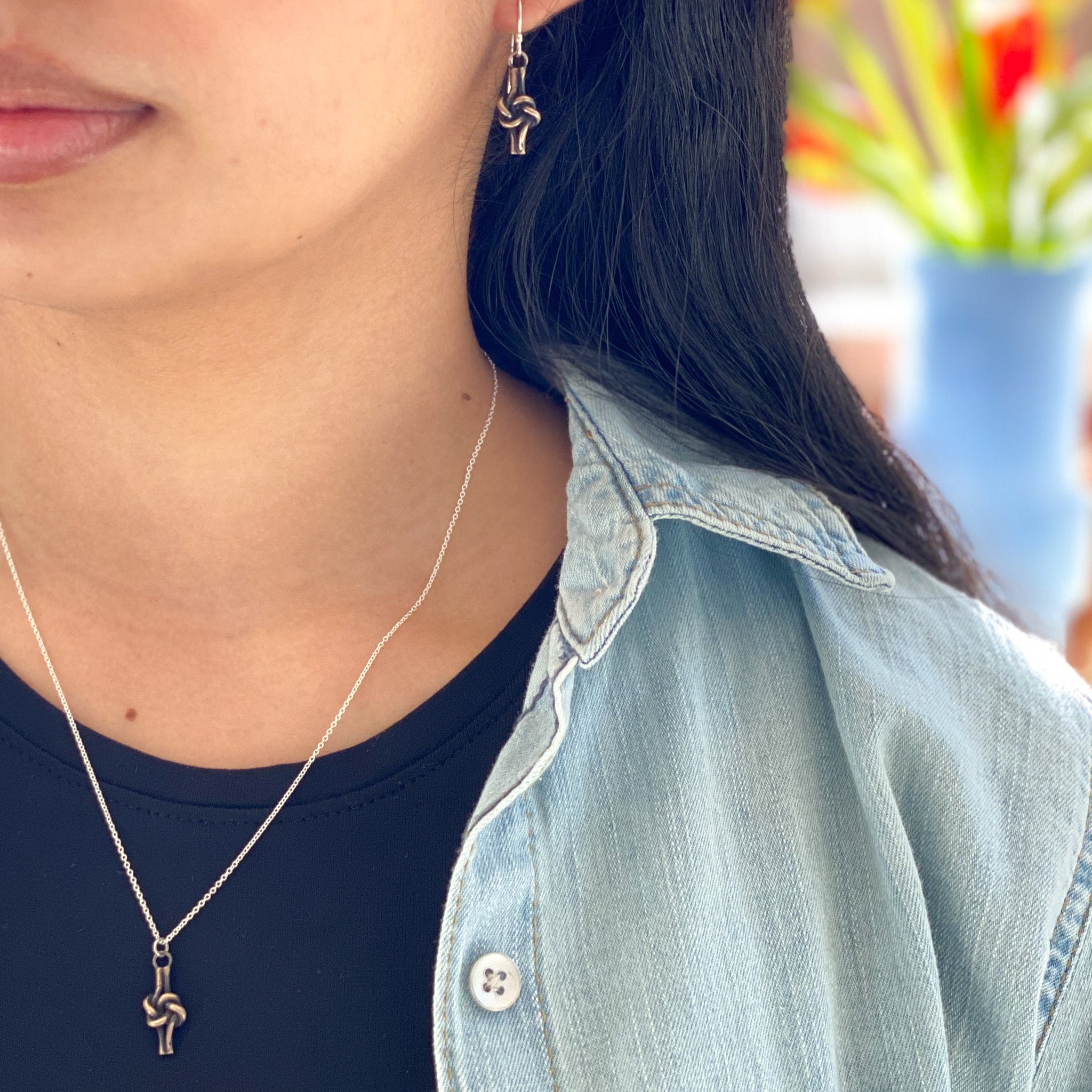 A close-up lifestyle shot of a person wearing a delicate silver chain necklace with a small bronze love knot pendant, along with matching earrings, a black t-shirt, and a light blue denim shirt.