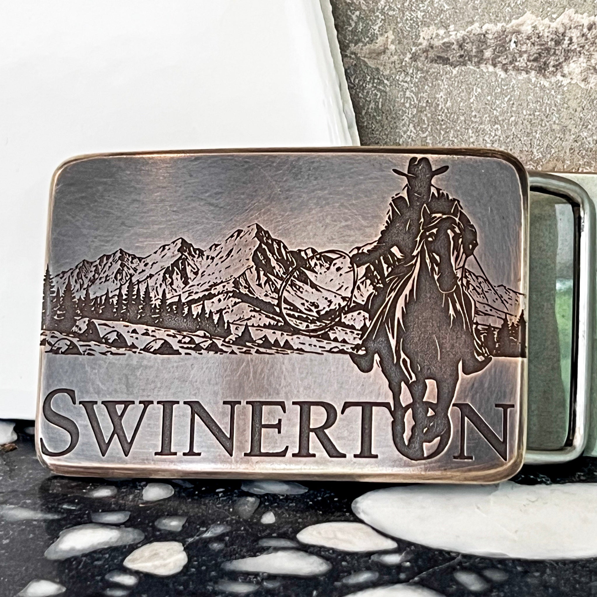 Bronze belt buckle with engraved design of a cowboy on a horse and mountains, branded 'Swinerton.