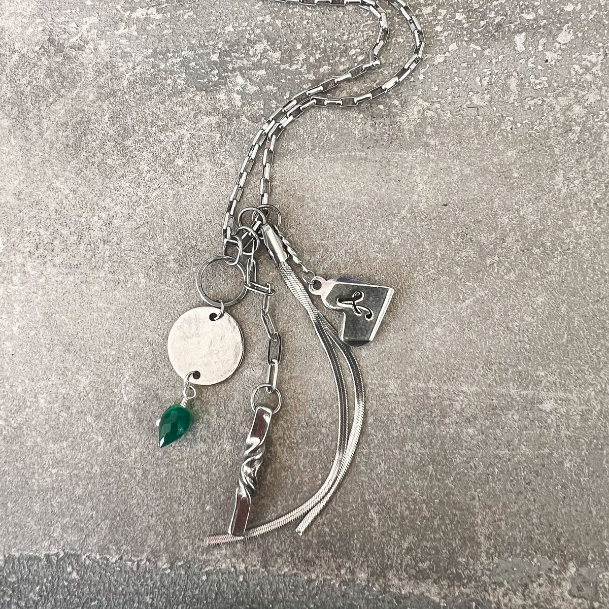 A silver-toned chain necklace with multiple charms, including a round disc, a small green teardrop pendant, a rectangular tag with a symbol, and curved ribbon-like elements, laid on a textured gray surface.