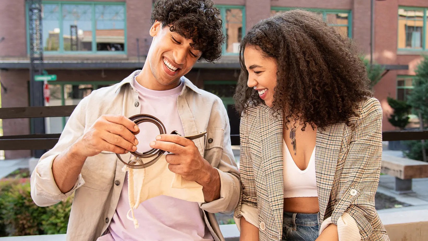Two young adults smiling and laughing outdoors while the man reads an engraving on the back of belt buckle and belt given to him.