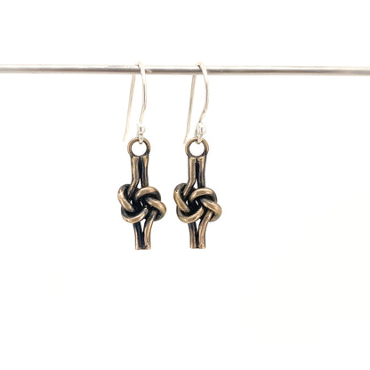 A pair of dangling earrings with sterling silver ear wires and  bronze love knot charms, hanging from a thin horizontal bar against a plain white background.