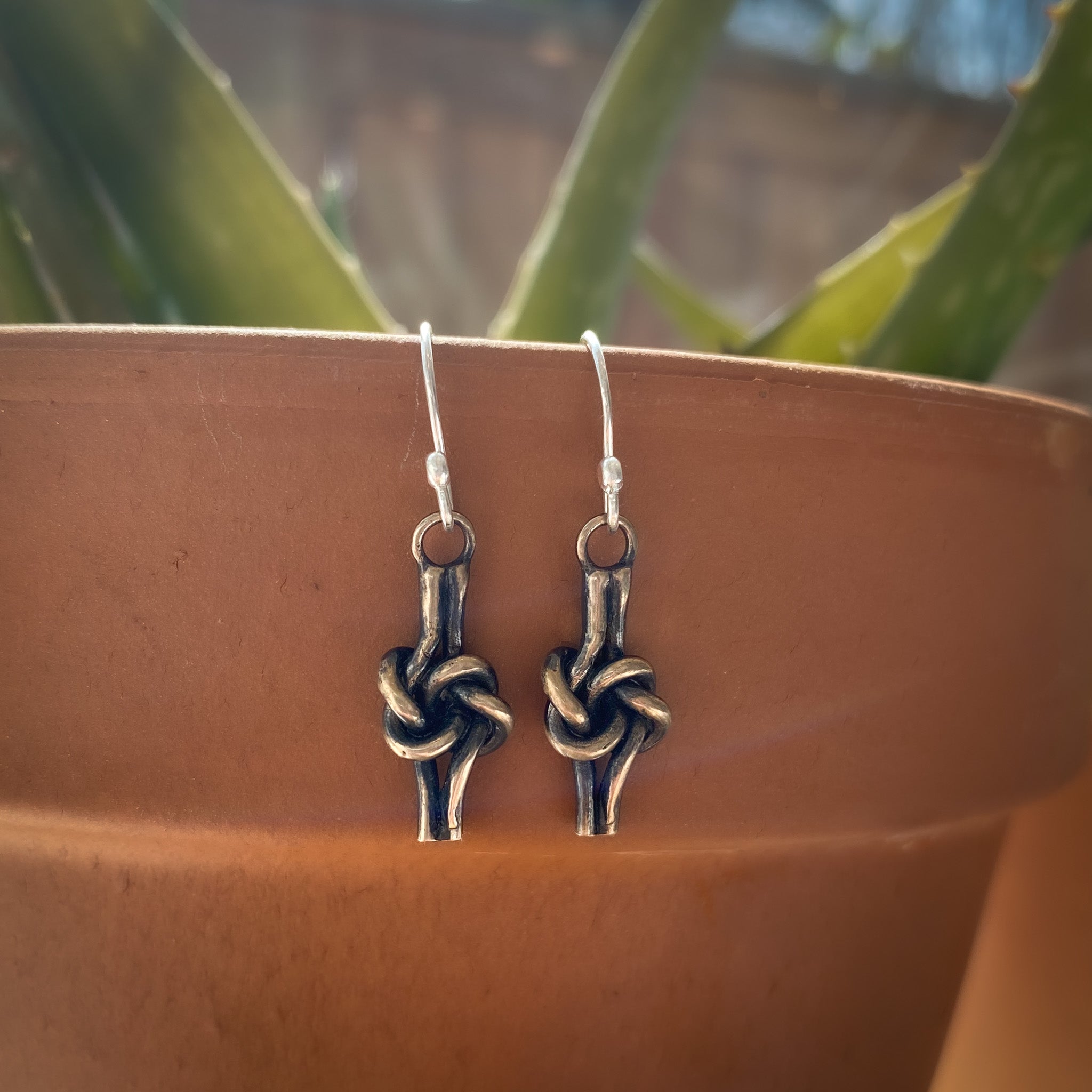 A pair of bronze love knot earrings with, resting on the rim of a terracotta pot with blurred green leaves in the background.