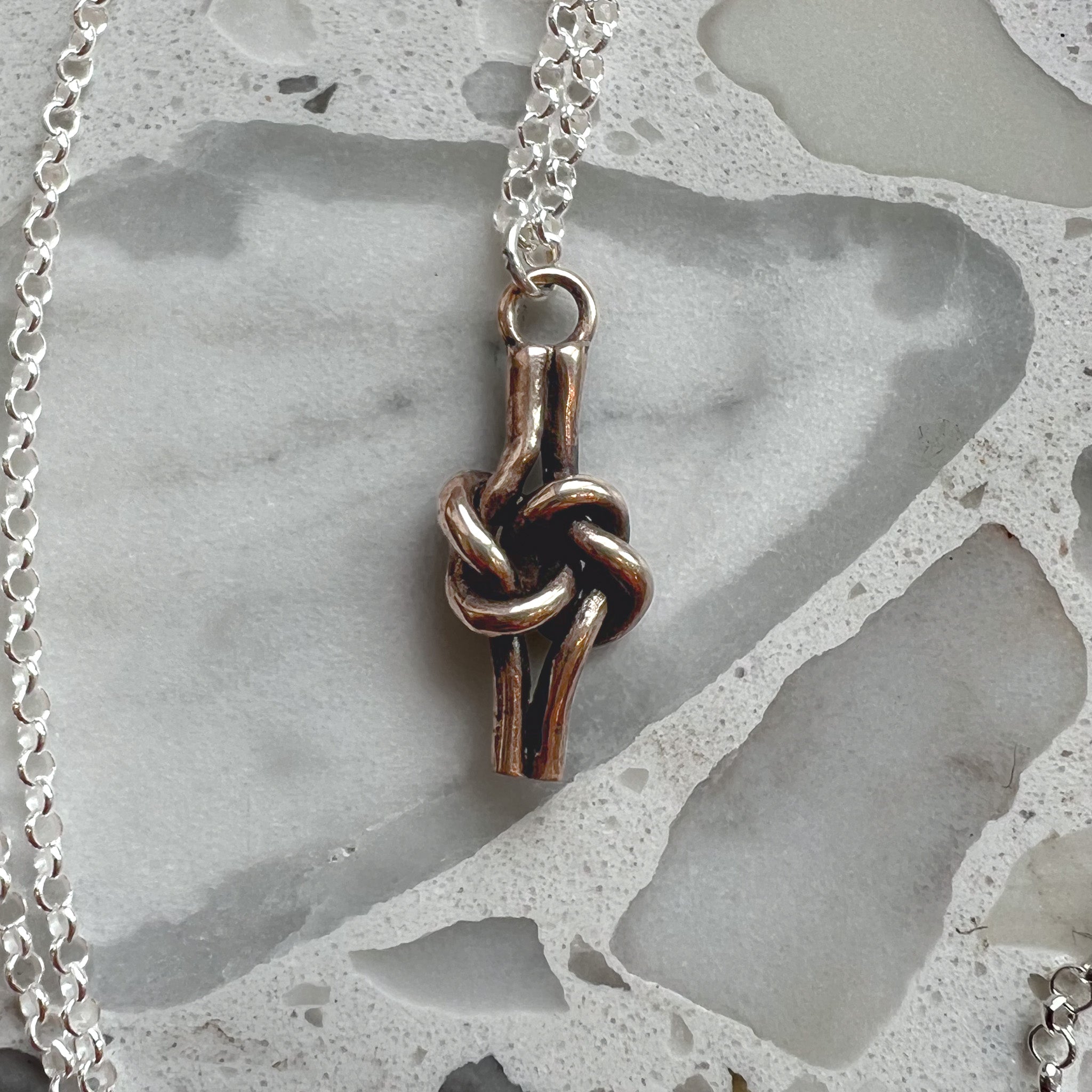 A close-up of a bronze love knot pendant hanging from a sterling silver chain on a light gray stone surface. Perfect for 8th anniversary celebration