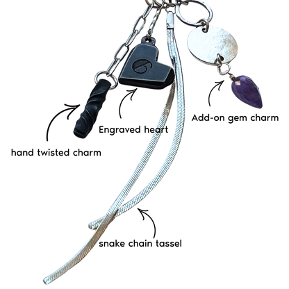Charms including an engraved heart, hand twisted charm, snake chain tassel, and add-on gem charm on a white background.