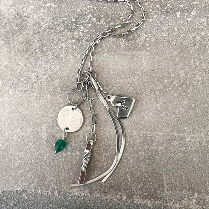A silver-toned chain necklace with multiple charms, including a round disc, a small green teardrop pendant, a rectangular tag with a symbol, and curved ribbon-like elements, laid on a textured gray surface.