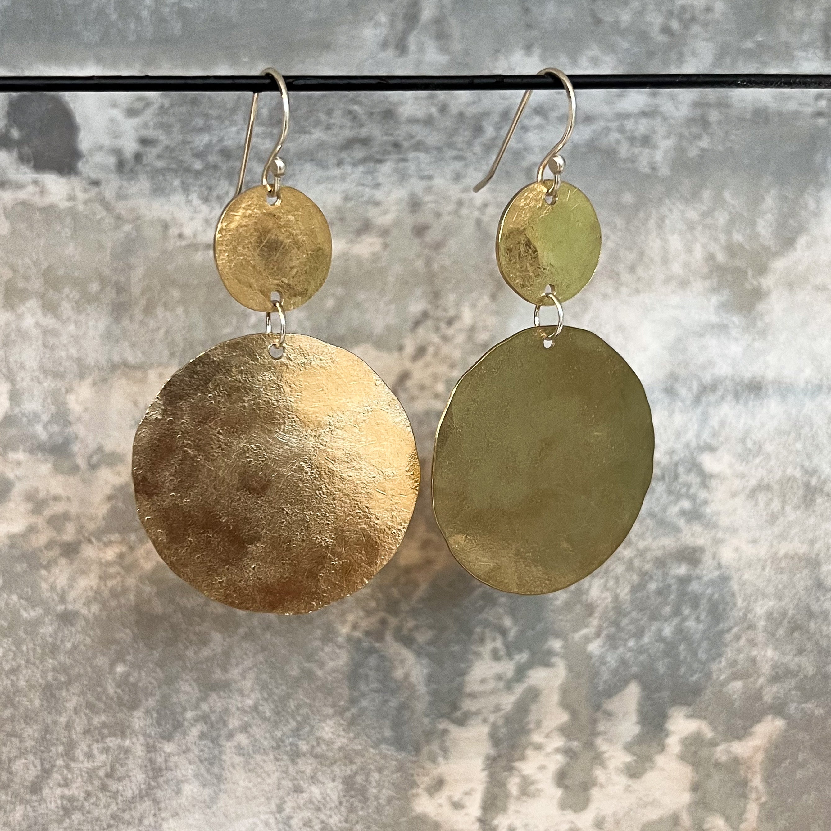 A pair of handmade dangle earrings with hammered circular discs in textured bronze, suspended from silver-toned fishhook wires against a mottled gray background.