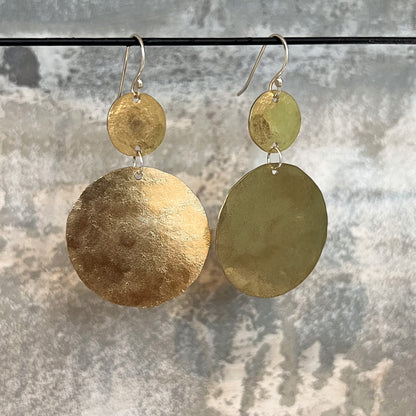 A pair of handmade dangle earrings with hammered circular discs in textured bronze, suspended from silver-toned fishhook wires against a mottled gray background.