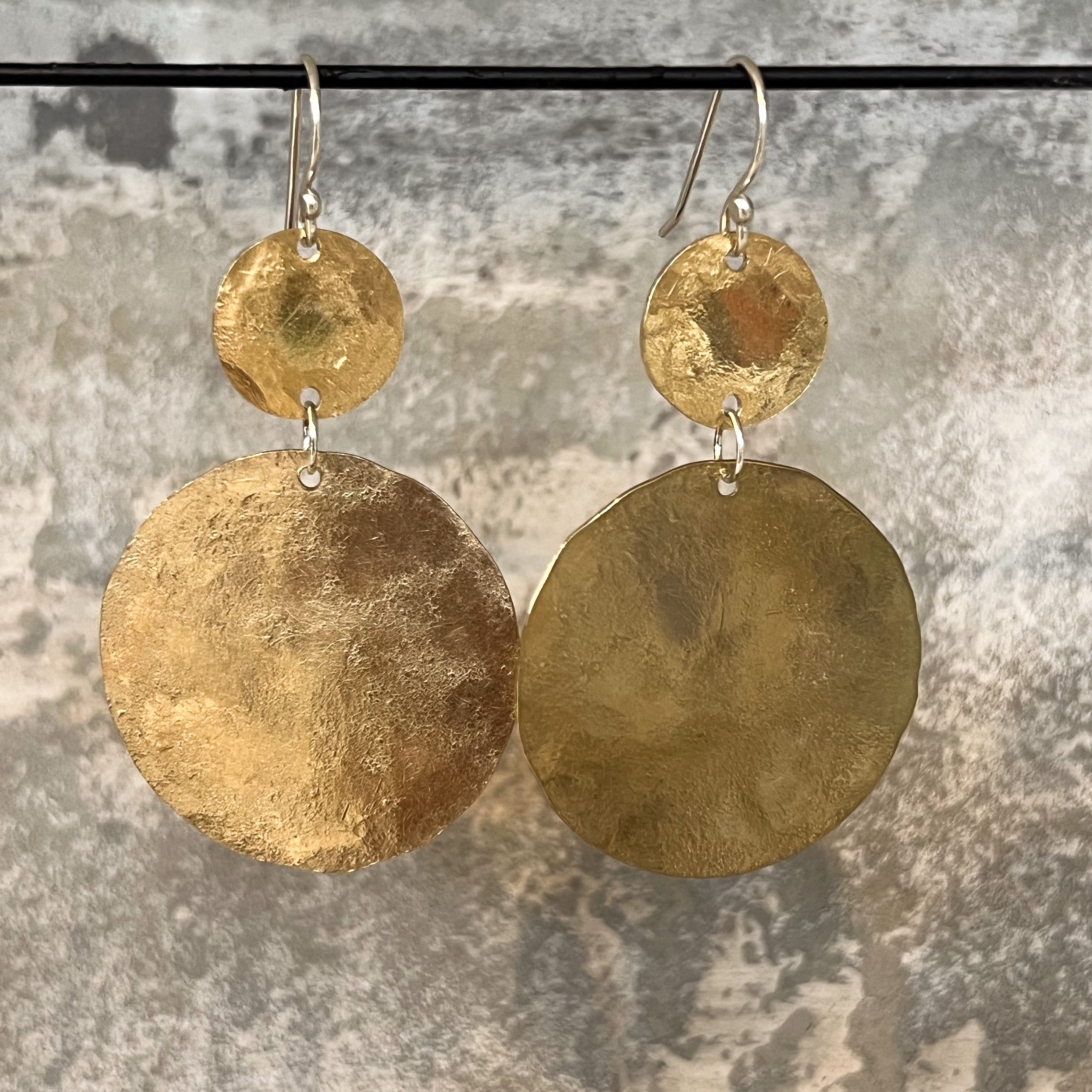 A pair of handmade dangle earrings with textured hammered yello bronze discs of different sizes and silver-tone fishhook wires, displayed against a mottled gray stone background.