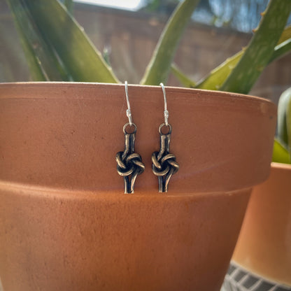 Bronze love knot earrings on planter edge with aloe leaves in background.